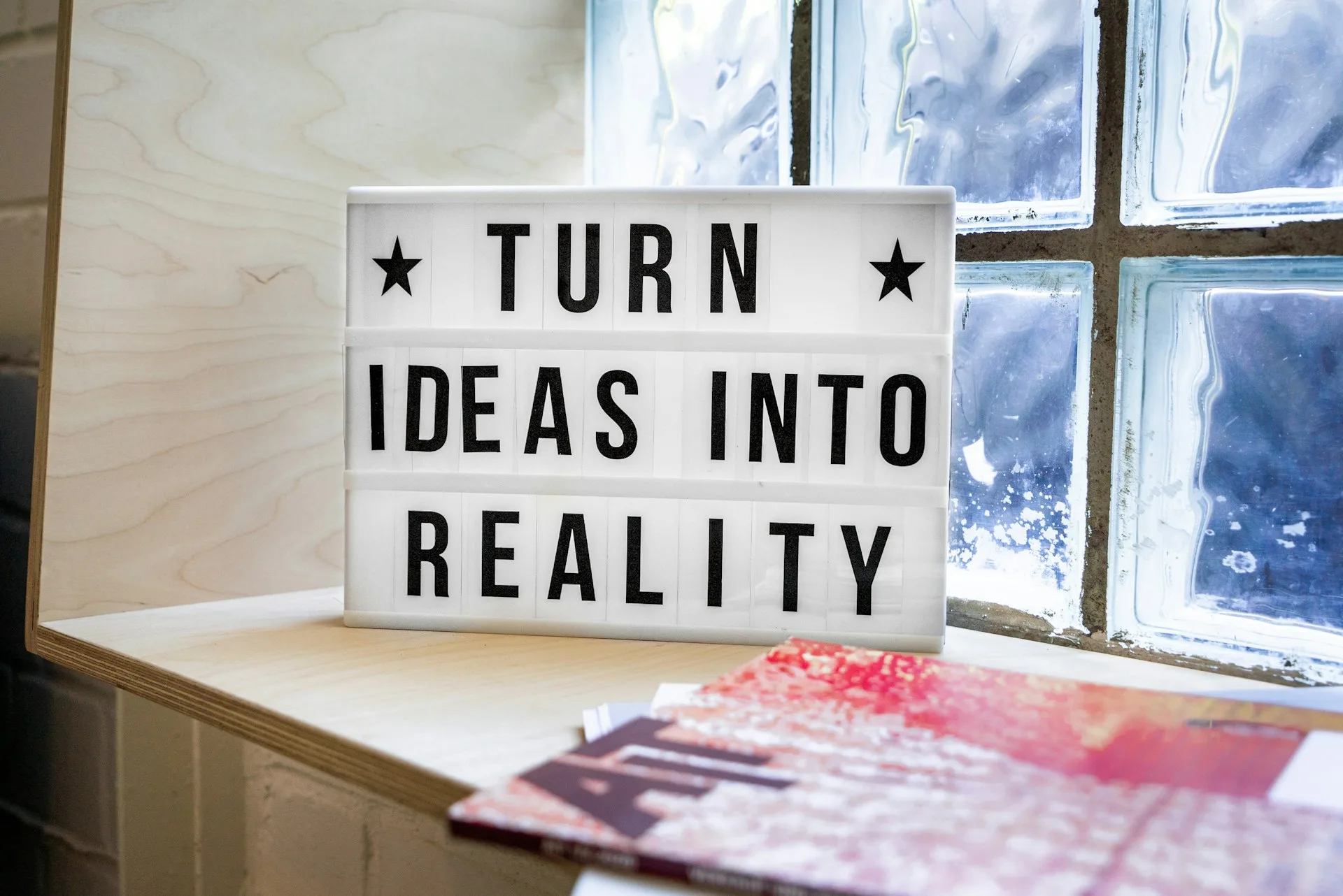 Inspirational lightbox sign with the message "Turn Ideas Into Reality," placed on a wooden shelf next to a frosted glass window and colorful magazines.